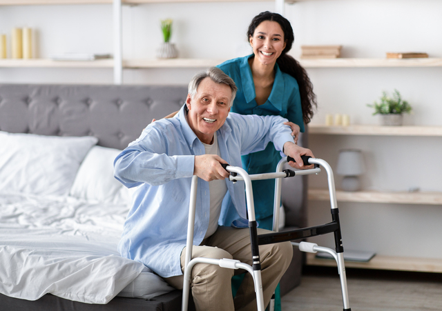 Positive female nurse helping elderly male with walking frame stand up from bed at home. Senior man receiving assistance from medical worker indoors. Professional care for disabled patients
